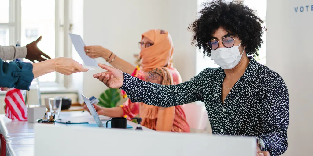 A woman wearing a mask passes someone a voting ballot from behind a desk