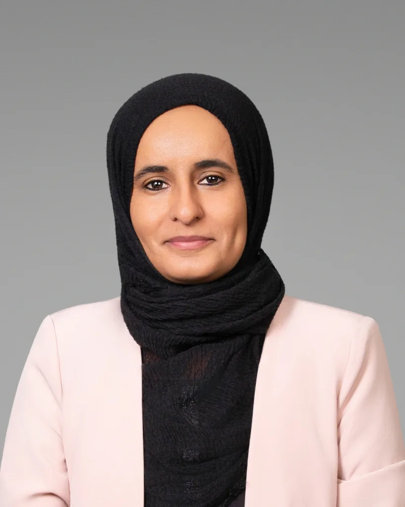 A woman wearing a black hijab and a light pink blazer is smiling slightly at the camera against a plain gray background.