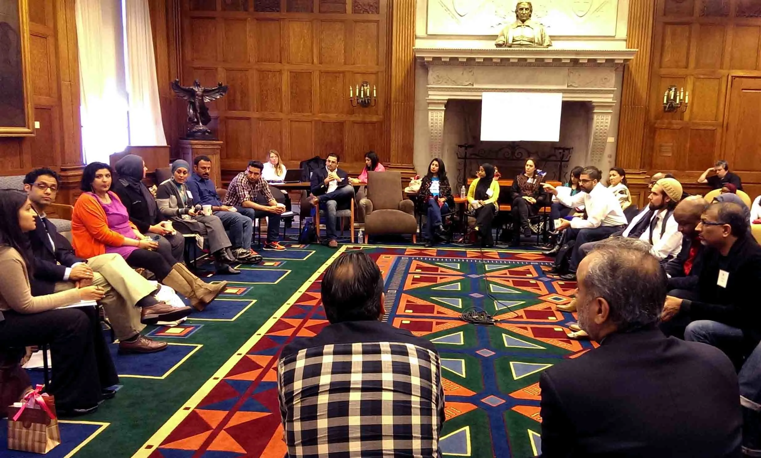 A diverse group of people sit in a large circle in a wood-paneled room with ornate decor, engaged in discussion. The room has a colorful geometric carpet and a fireplace with a statue above it.