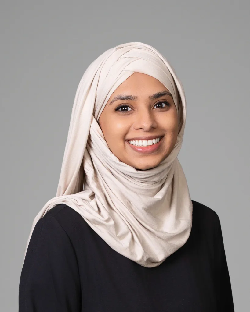 A woman wearing a light beige hijab and a black top smiles at the camera against a plain gray background.