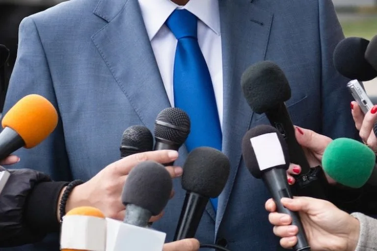 A man in a suit is surrounded by reporters with microphones