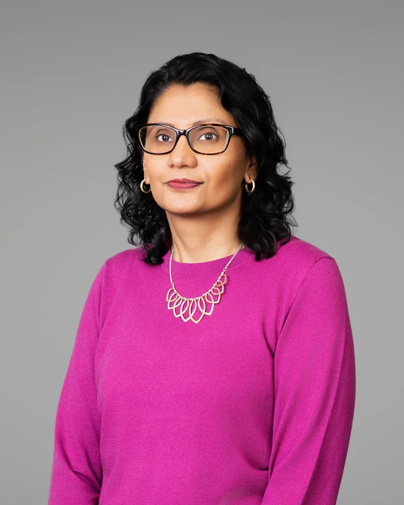 A woman with shoulder-length curly black hair and glasses wears a bright pink sweater and a gold necklace, standing against a plain gray background.