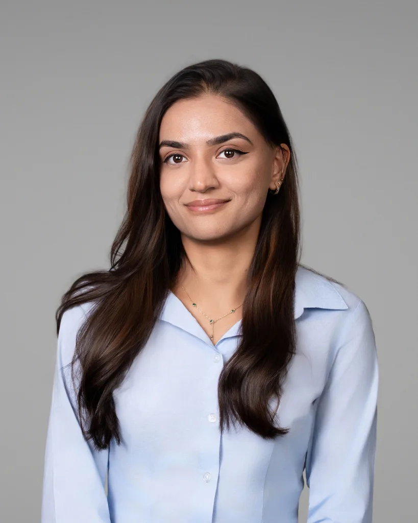 A woman with long brown hair wearing a light blue collared shirt is smiling softly. She is posed against a plain grey background.