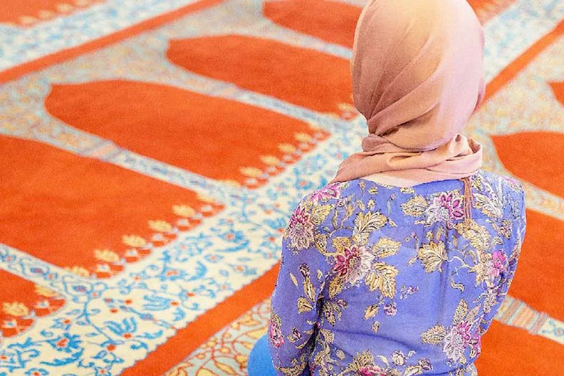 A person wearing a light pink headscarf and a purple floral top is sitting on an ornate red and blue patterned prayer rug, viewed from behind.