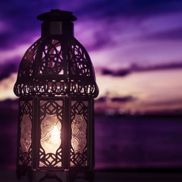 Ornate lantern with intricate patterns glowing softly at dusk, set against a blurred background of a purple and blue sunset sky.