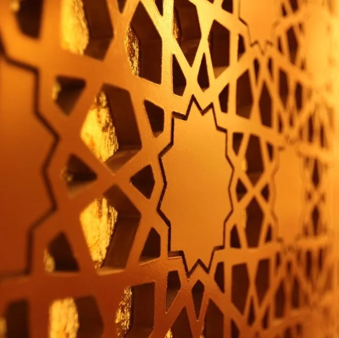 Close-up of an intricate geometric pattern, resembling traditional Islamic art, with star-shaped cutouts in a warm golden light, creating depth and shadow on the textured surface.