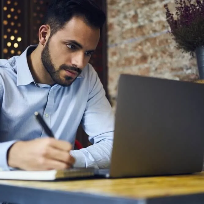 A man with a beard, wearing a light blue shirt, sits at a desk writing in a notebook while looking at a laptop. The setting appears to be a modern, cozy workspace with a brick wall in the background.