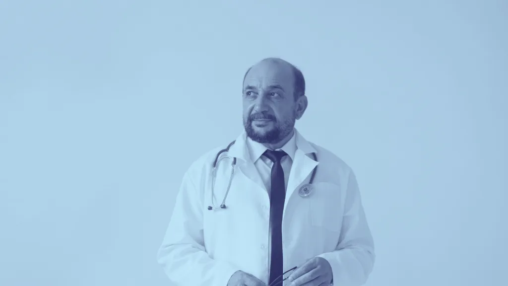A middle-aged male doctor wearing a white coat, stethoscope, and tie stands against a plain light blue background, looking thoughtfully to the side.