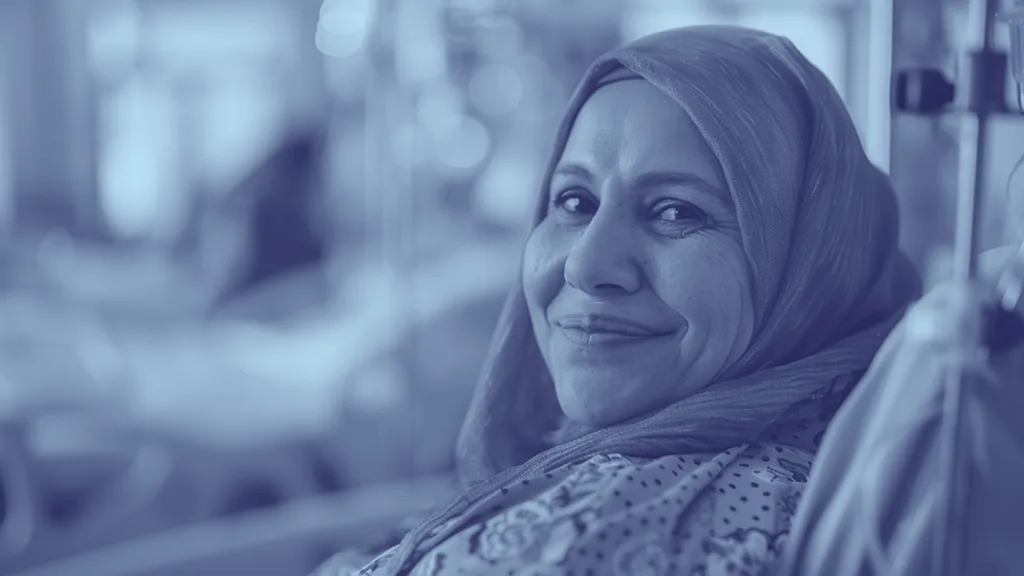 A woman wearing a patterned hijab smiles warmly while sitting in what appears to be a medical or hospital setting. The image has a blue filter.