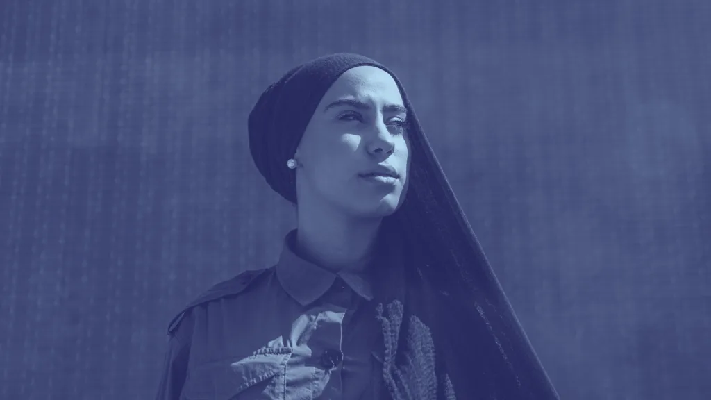 A woman wearing a headscarf and buttoned shirt looks confidently into the distance, standing against a textured wall. The image has a blue monochrome filter.