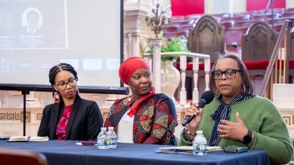 Three women sit at a table during a panel discussion. One woman, wearing glasses and a green sweater, speaks into a microphone while the others listen. Notebooks and water bottles are on the table in front of them.