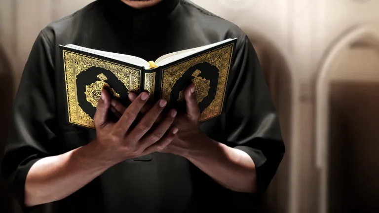 A person wearing a black garment holds and reads an open Quran with an ornate gold cover, standing indoors against a soft, blurred background.