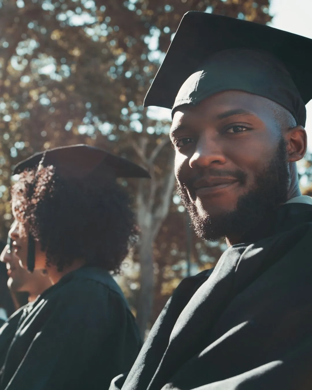A man wearing a graduation cap and gown smiles at the camera while sitting outdoors. Another person in a cap and gown is visible beside him. Trees and sunlight can be seen in the background.