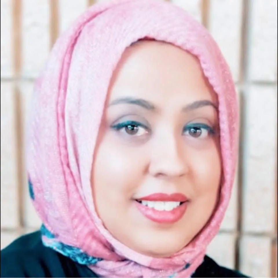 A woman wearing a pink hijab smiles at the camera. The background consists of beige, rectangular bricks.