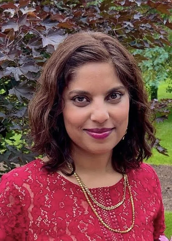 A woman with wavy brown hair wearing a red lace top and gold necklace stands outdoors in front of leafy green and purple foliage, smiling at the camera.