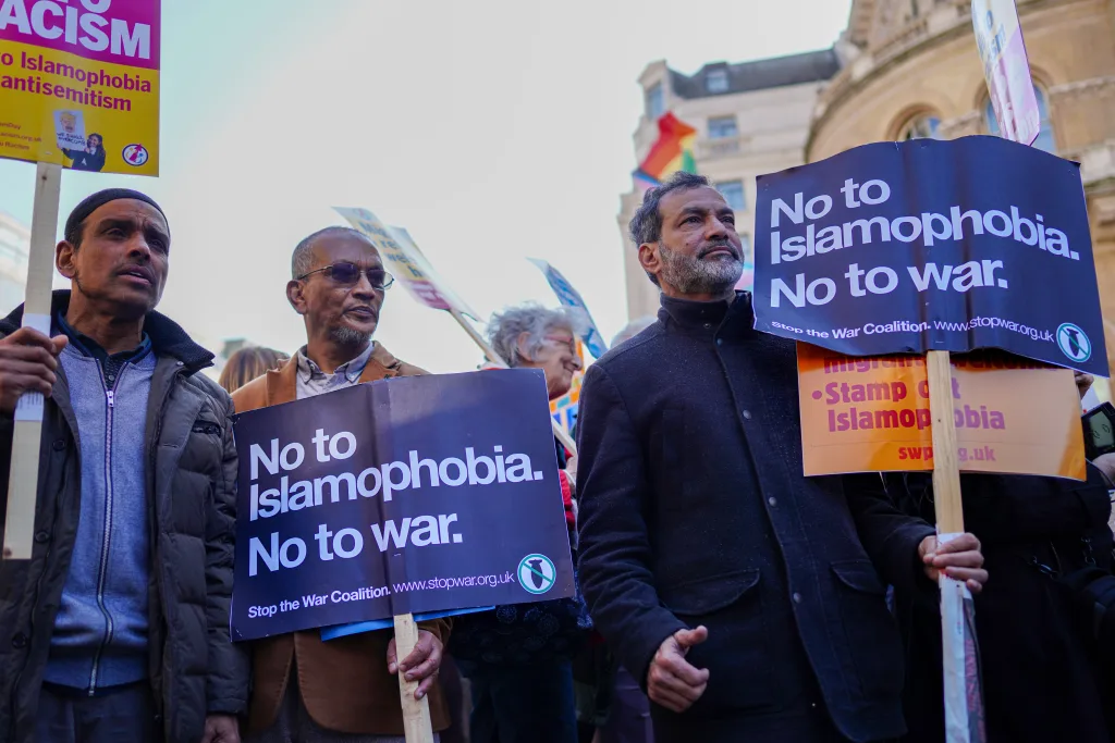 A group of people at a protest hold signs reading “No to Islamophobia. No to war.” and “Stamp out Islamophobia.” A rainbow flag and other protest signs are visible in the background.