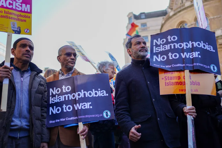 A group of people at a protest hold signs reading “No to Islamophobia. No to war.” and “Stamp out Islamophobia.” A rainbow flag and other protest signs are visible in the background.