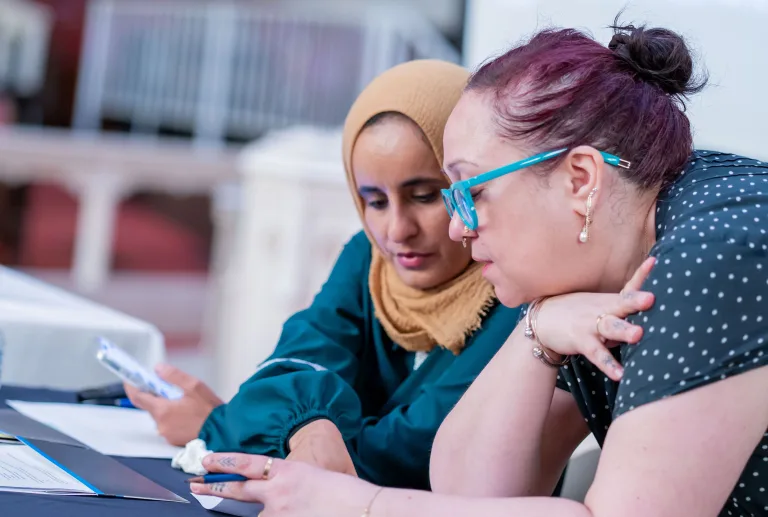 Two women sit at a table, closely looking at documents and phones together. One wears a tan hijab, while the other has glasses and a black shirt. They appear to be discussing or reviewing information.