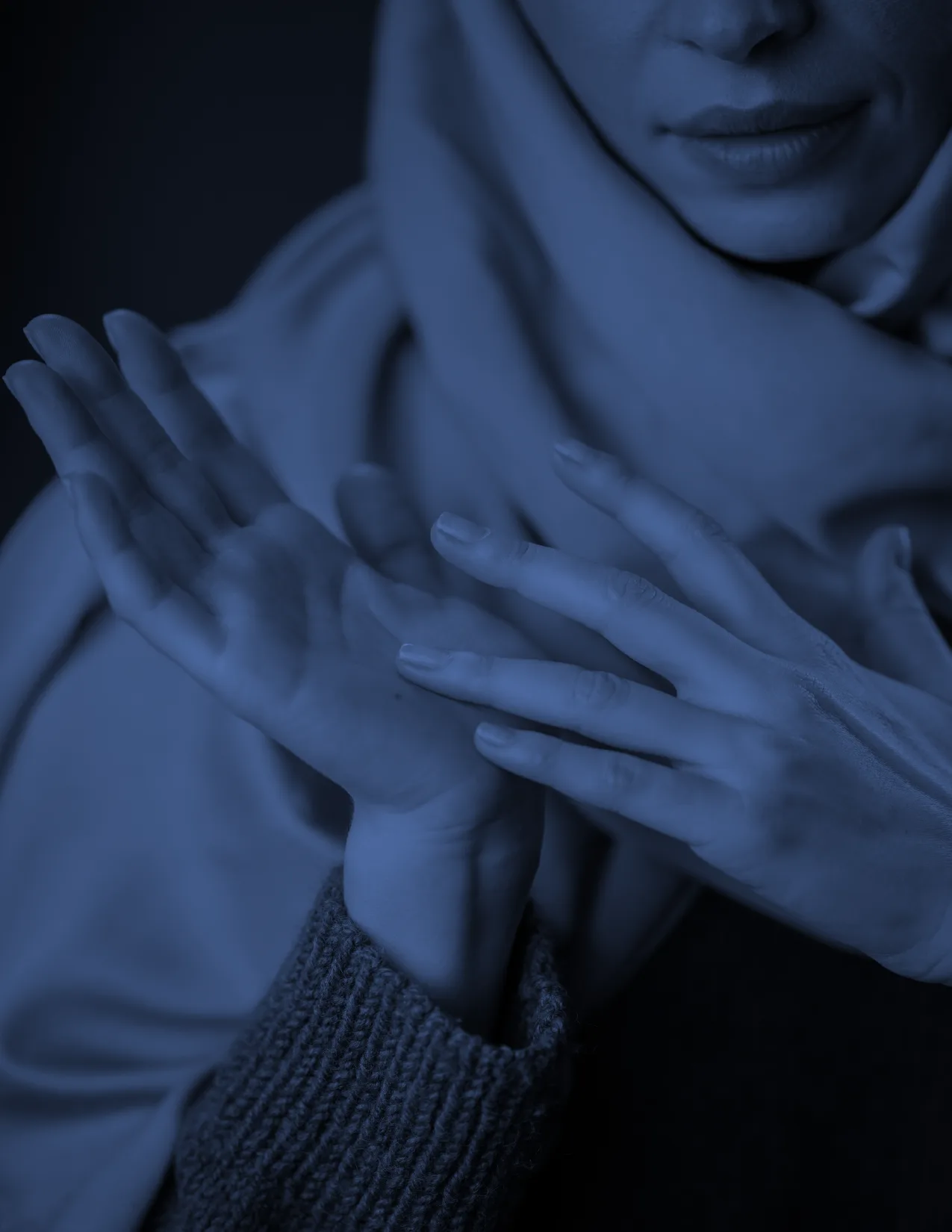 A person in a headscarf is shown from the shoulders up, holding and gently touching their wrist with their other hand. The image has a blue tint and a soft focus.
