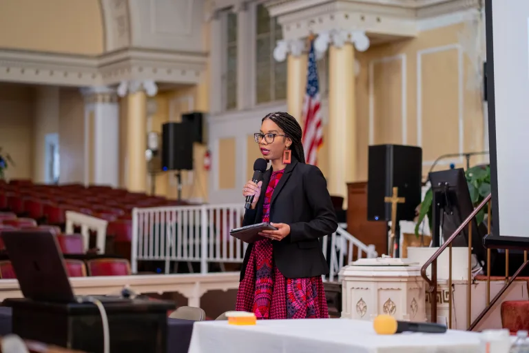 A woman wearing glasses and a patterned dress speaks into a microphone while holding a tablet, standing in a church or auditorium with rows of empty red seats and an American flag in the background.