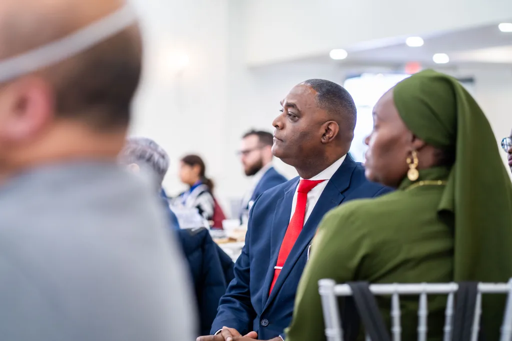 A man in a suit and red tie sits attentively next to a woman wearing a green hijab at a formal indoor event, with other attendees visible in the background.