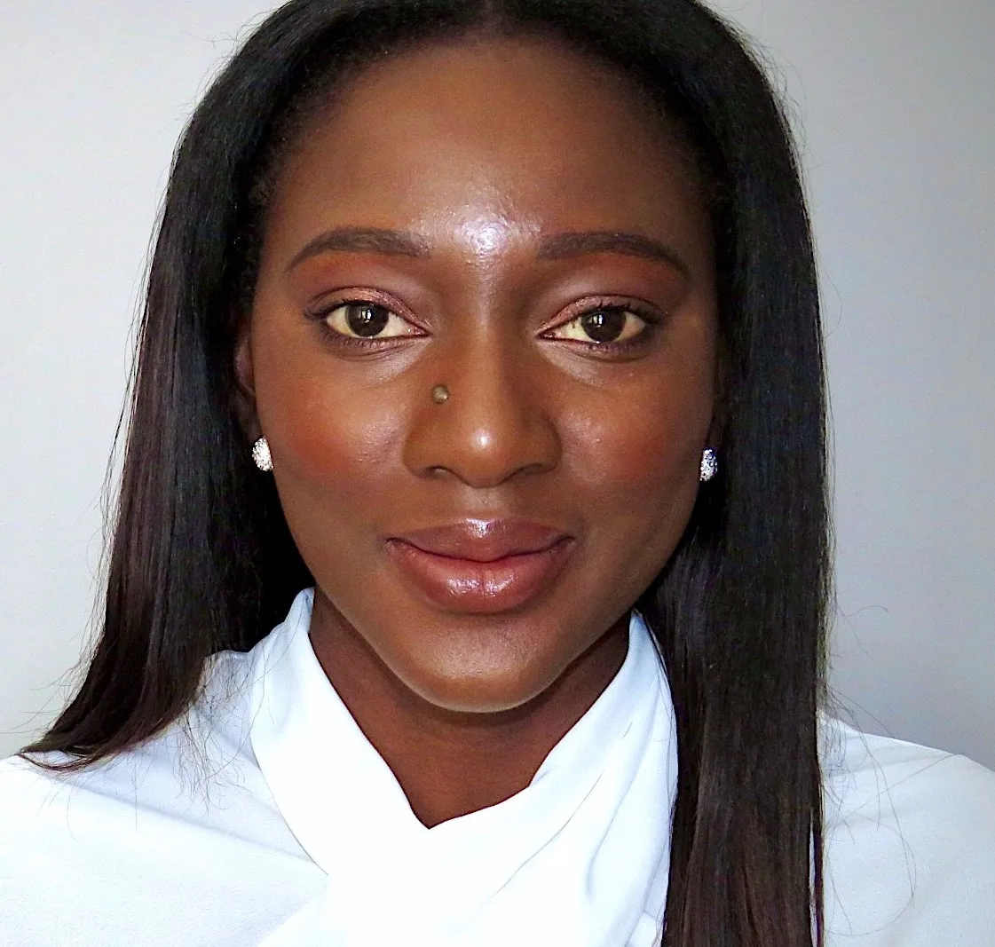 A woman with long straight hair, wearing a white blouse and stud earrings, smiles gently at the camera against a plain light background.