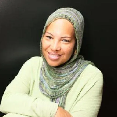 A woman wearing a light green hijab and shirt smiles warmly with her arms crossed in front of a plain black background.