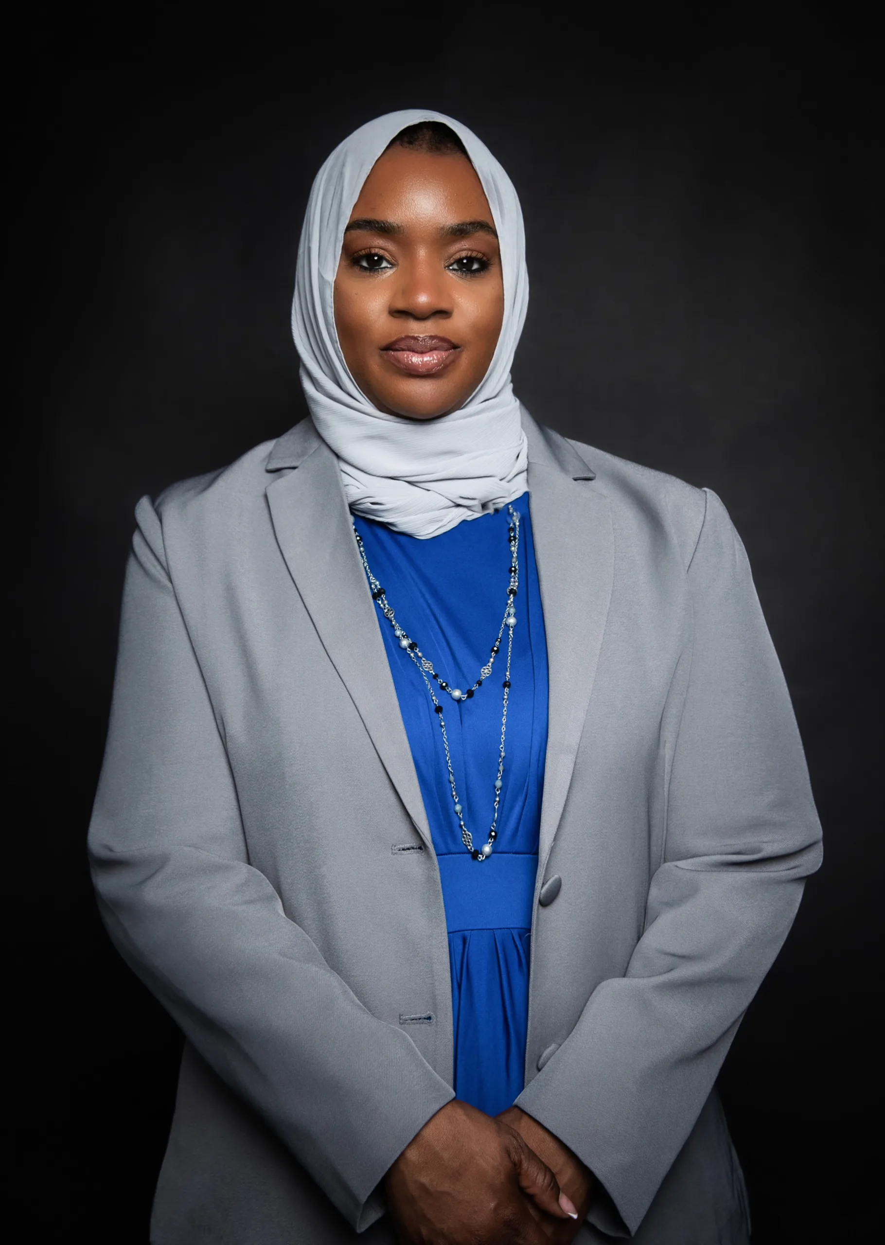 A woman wearing a light gray hijab and blazer over a blue dress stands against a dark background, facing the camera with a calm expression, hands folded in front.