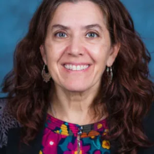 Woman with long wavy brown hair wearing hoop earrings, a colorful patterned blouse, and a dark blazer, smiling in front of a blue background.