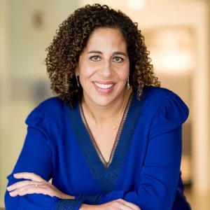 Woman with curly hair wearing a royal blue blouse, smiling and resting her arms on a surface, with a blurred indoor background.