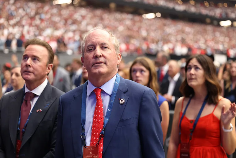 A man in a blue suit with a red tie stands in a crowded arena, surrounded by other formally dressed people, with a blurred audience in the background.
