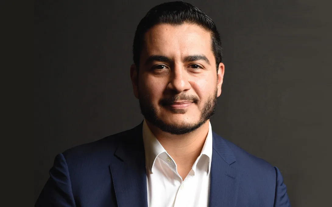A man with short dark hair and a trimmed beard is wearing a navy blue suit jacket and white shirt, posing against a plain dark background, looking directly at the camera with a slight smile.