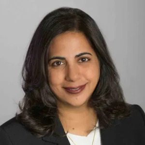 A woman with shoulder-length dark hair, wearing a black blazer and white top, smiles at the camera against a plain light gray background.